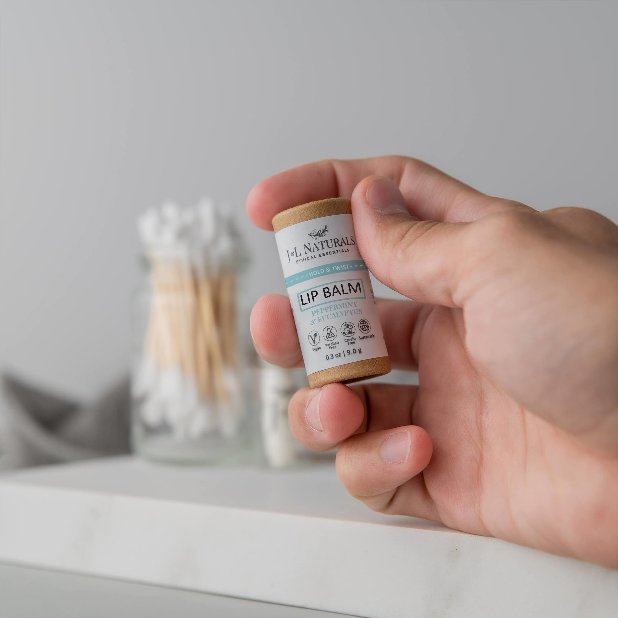 A hand holding a lip balm stick against a modern bathroom counter setup.