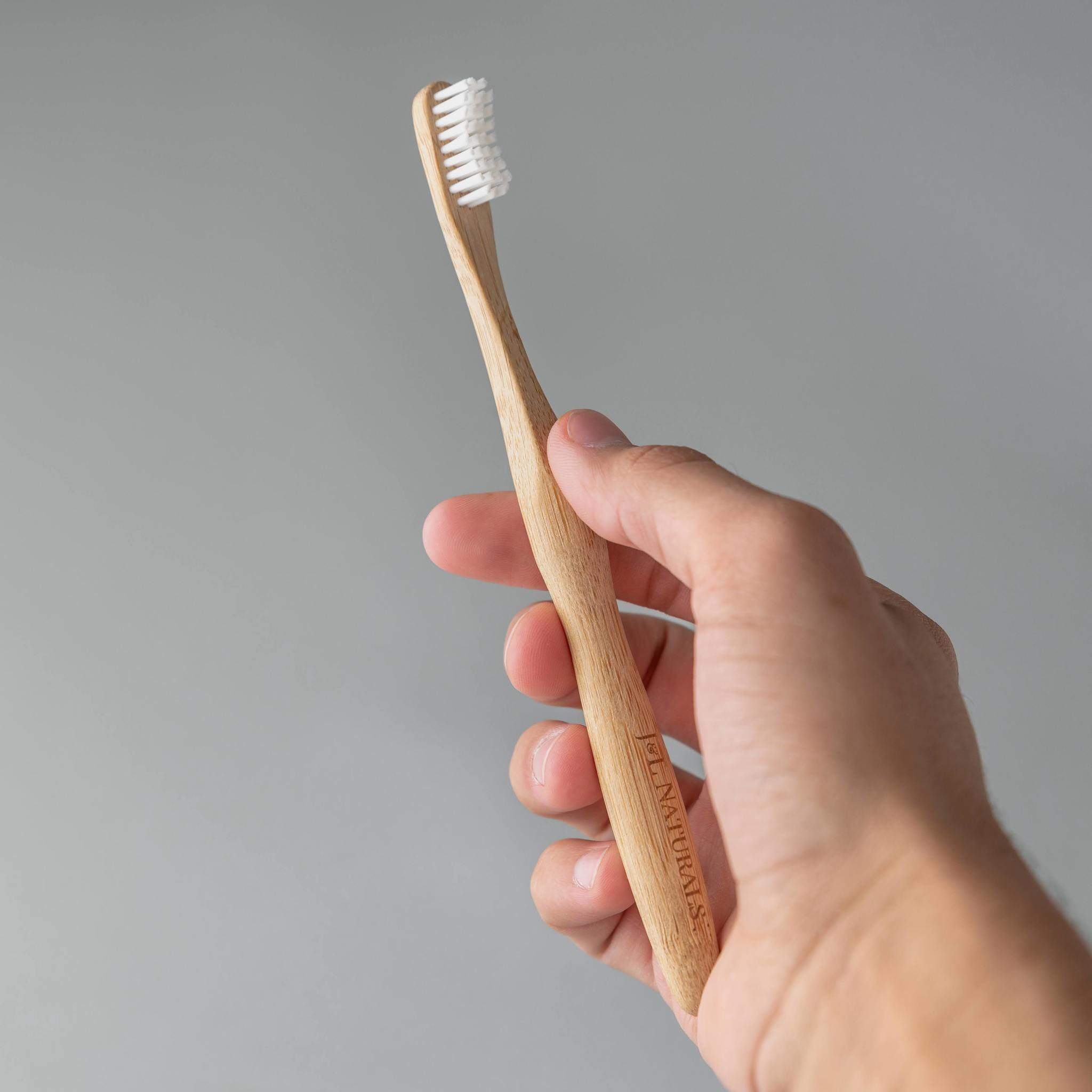 A hand holding a bamboo toothbrush with white bristles against a neutral background.