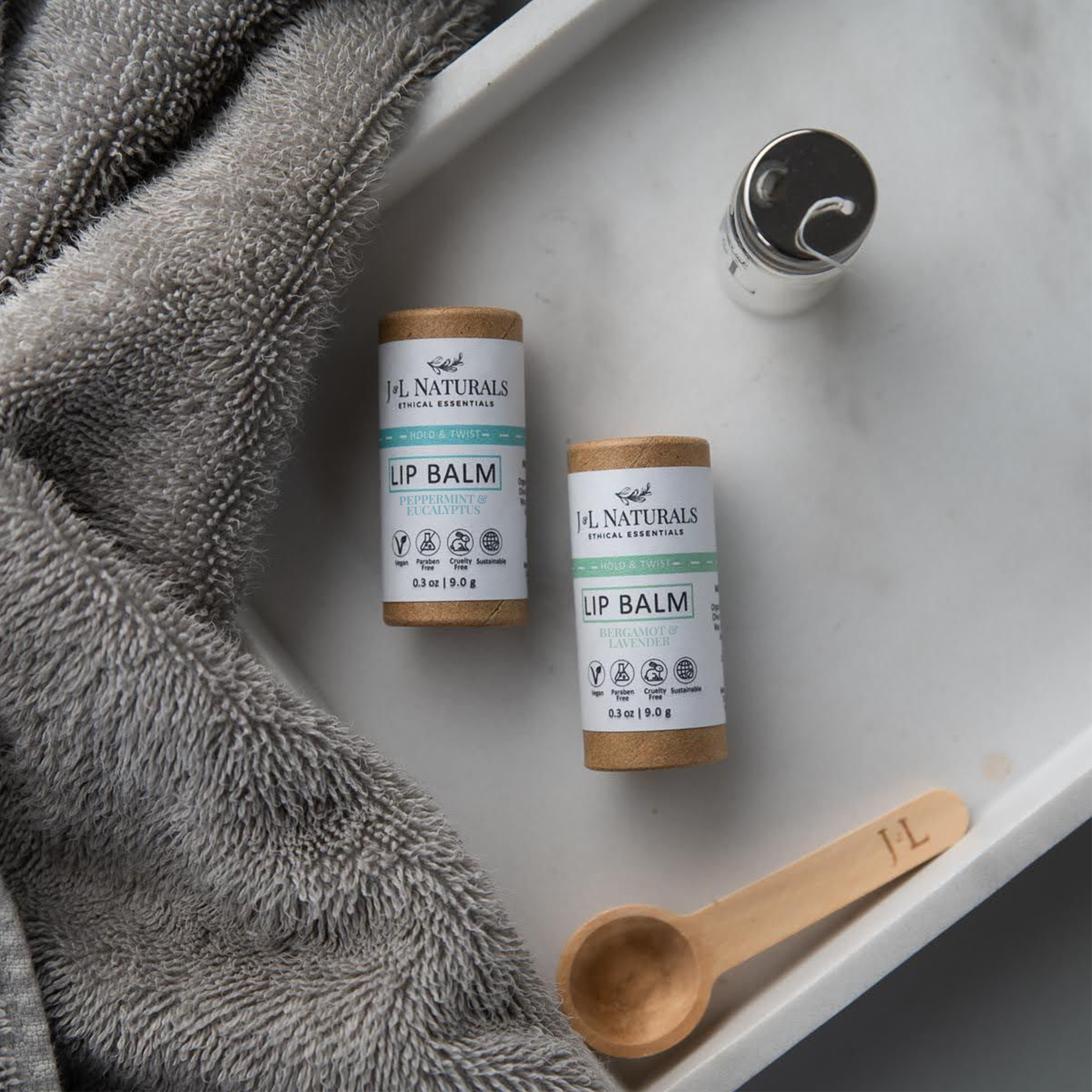 Two eco-friendly lip balm sticks placed on a tray with a soft gray blanket, wooden spoon, and minimal decor.
