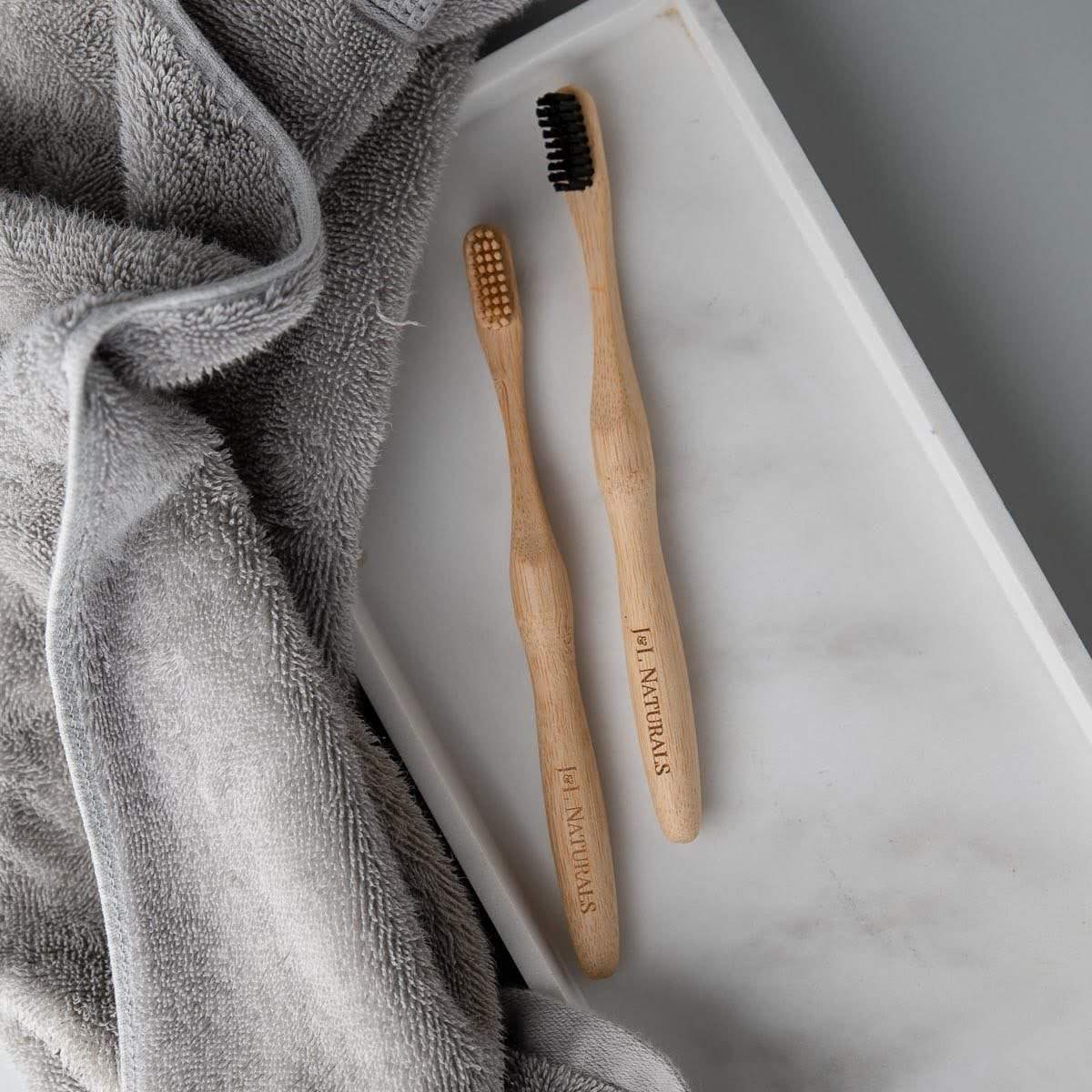 Two bamboo toothbrushes with black and natural bristles placed on a soft gray towel and white countertop.