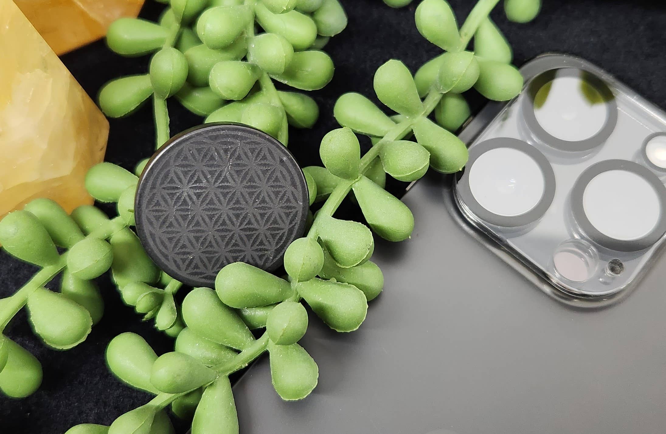 Shungite EMF Protection Tile attached to the back of a smartphone, resting on a table with a green plant nearby, blending functionality with natural aesthetics.