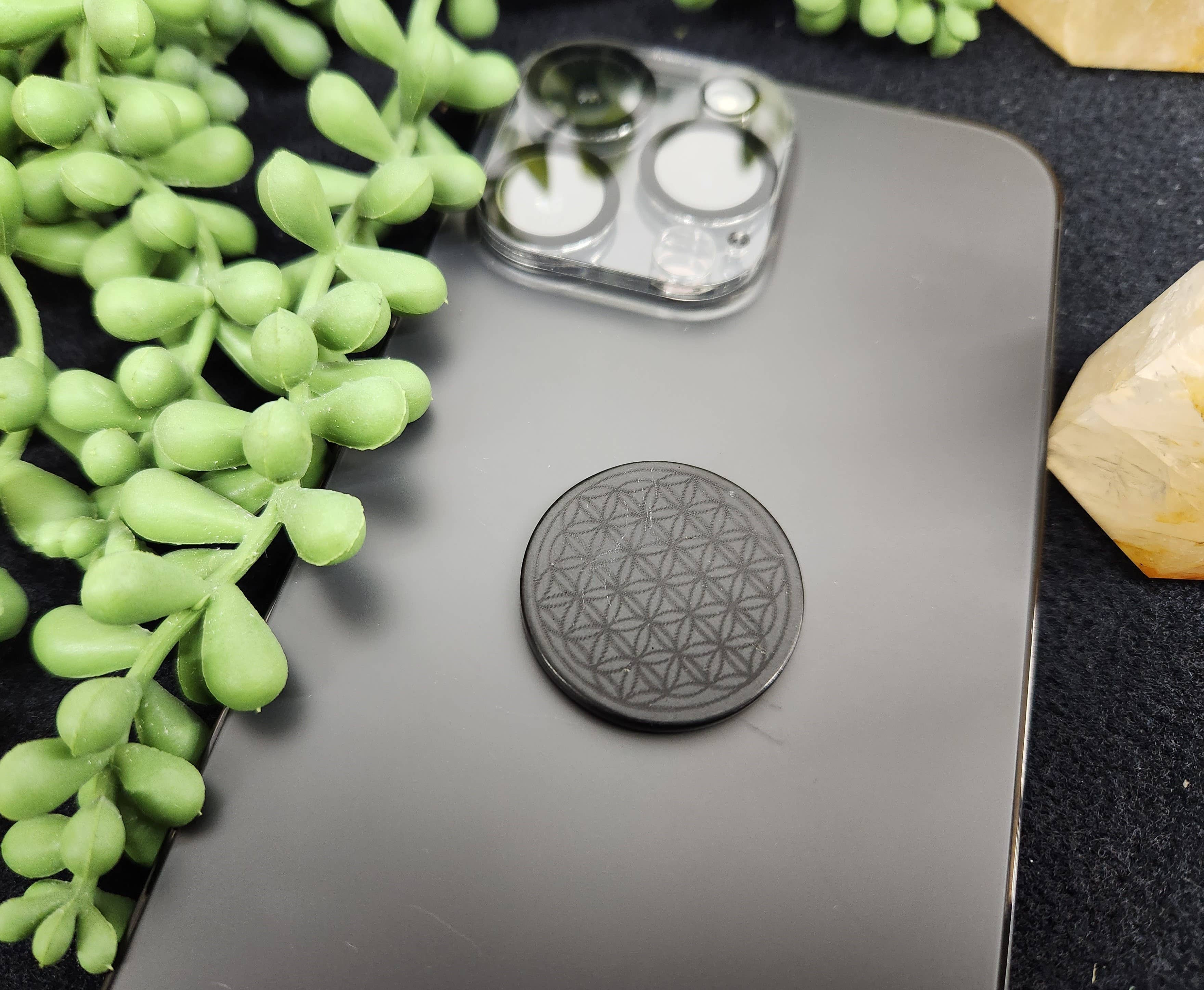 Shungite EMF Protection Tile attached to the back of a smartphone, resting on a table with a green plant nearby, blending functionality with natural aesthetics.