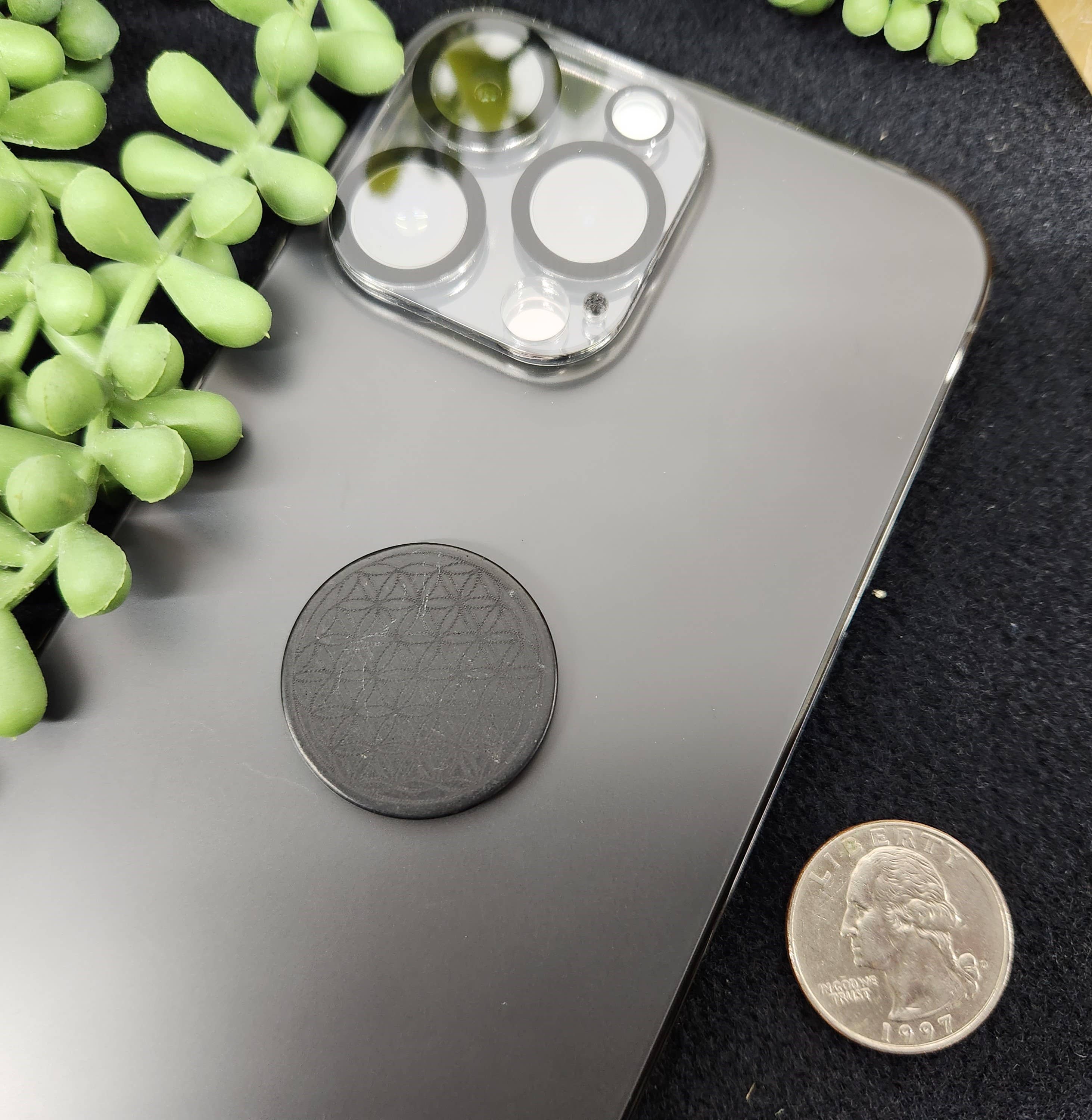 Shungite EMF Protection Tile attached to the back of a smartphone, resting on a table with a green plant nearby, blending functionality with natural aesthetics.