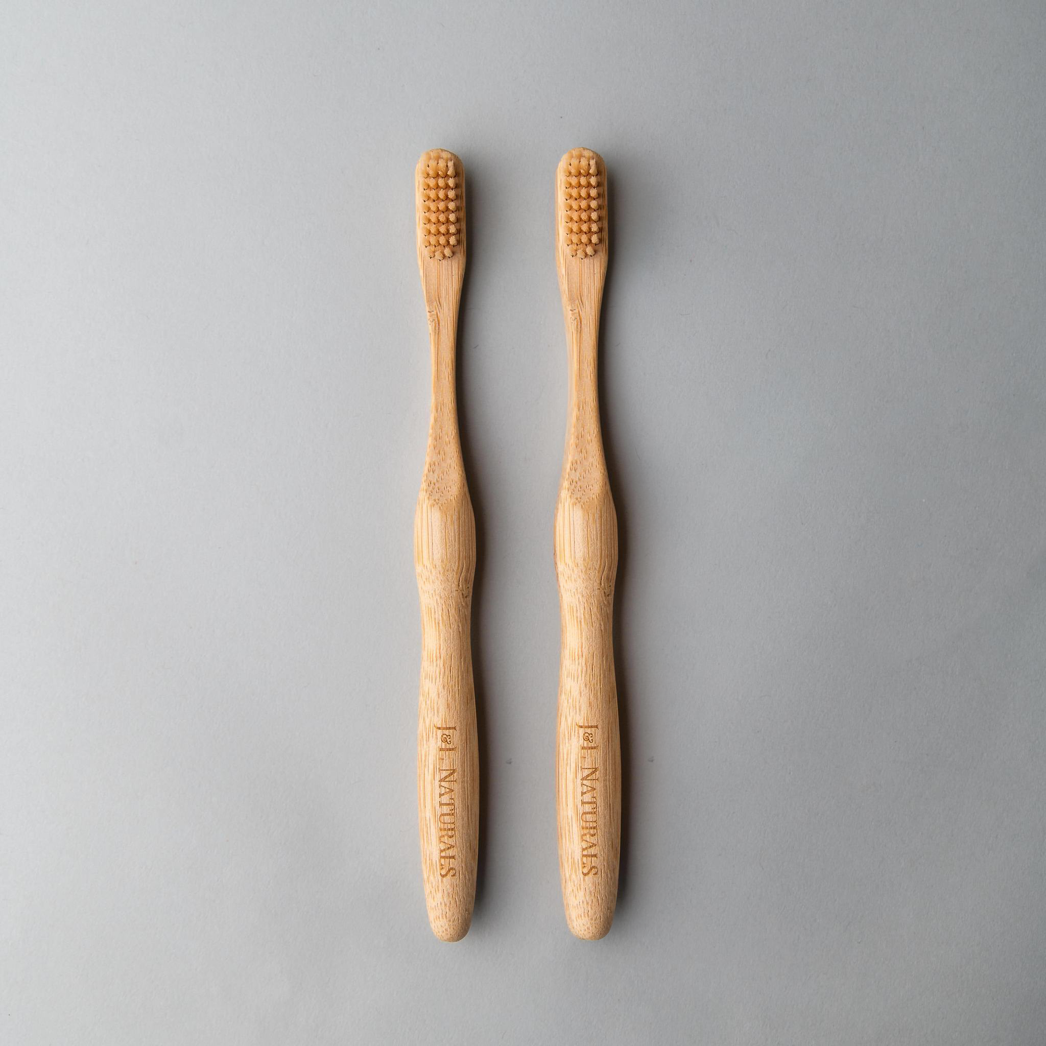 Two bamboo toothbrushes with natural/tan bristles, arranged side by side on a gray background.