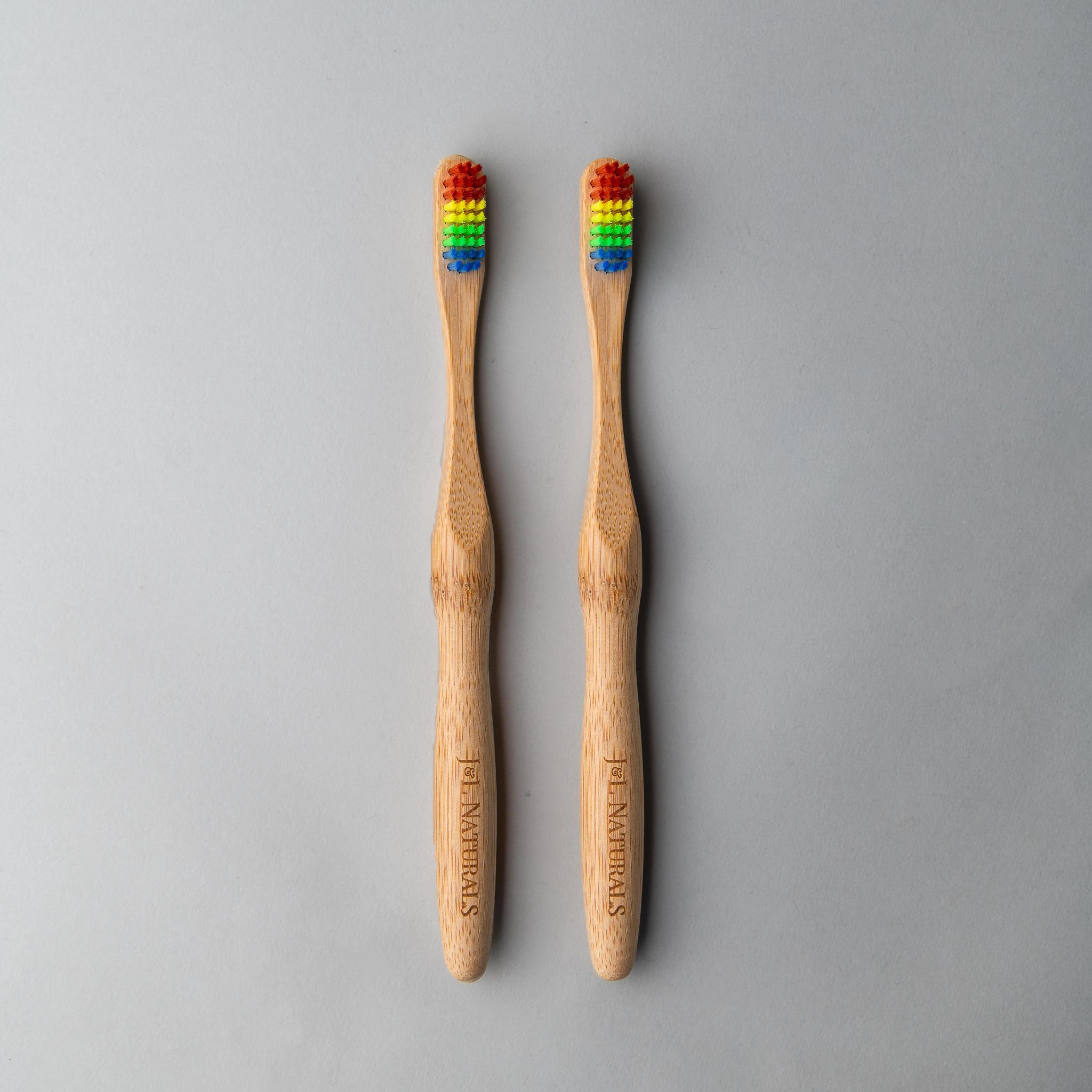 Two bamboo toothbrushes with rainbow bristles, arranged side by side on a gray background.