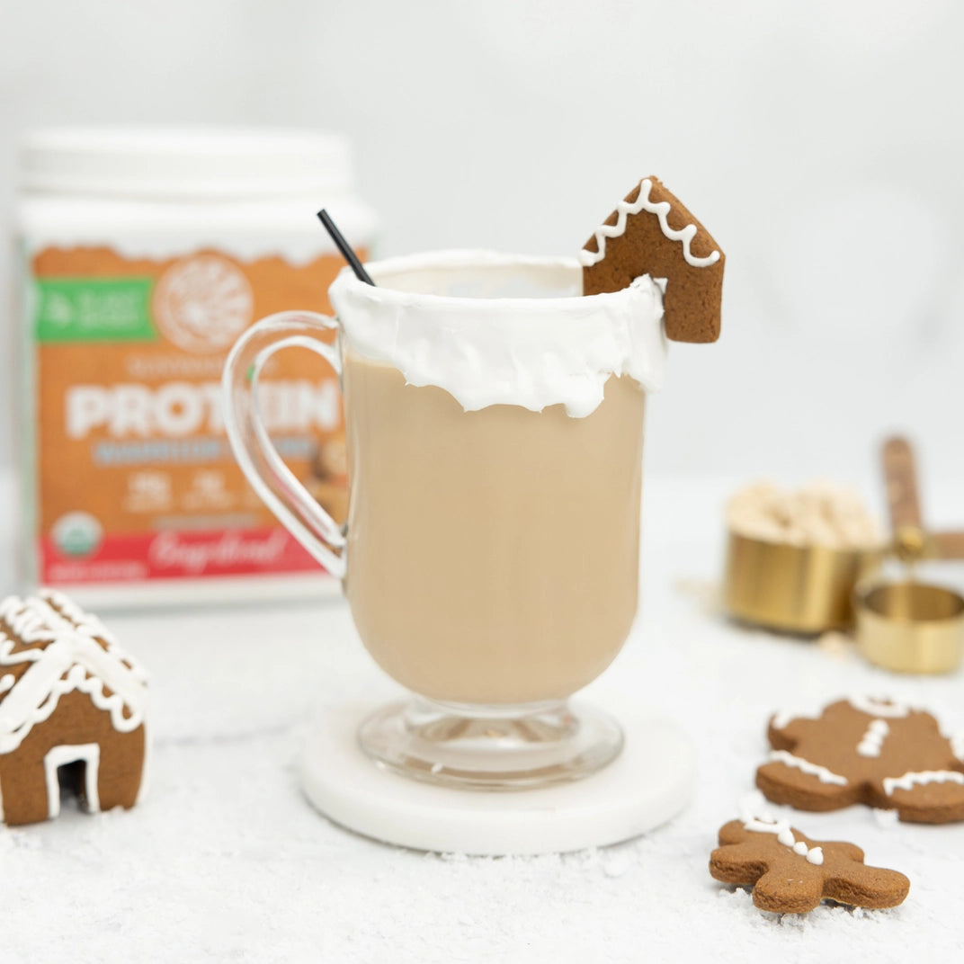 mug with gingerbread house cookie on the rim, protein powdeer in background, gingerbread house on left and gingerbread man cookie broken on right