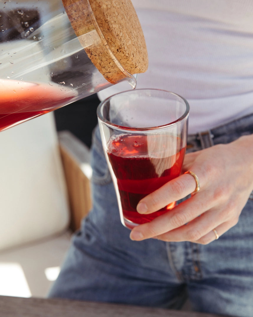 hand holding cup being filled from a glass pitcher, cup is half full of red liquid a person with jeans and white t-shirt are holding both in a kitchen