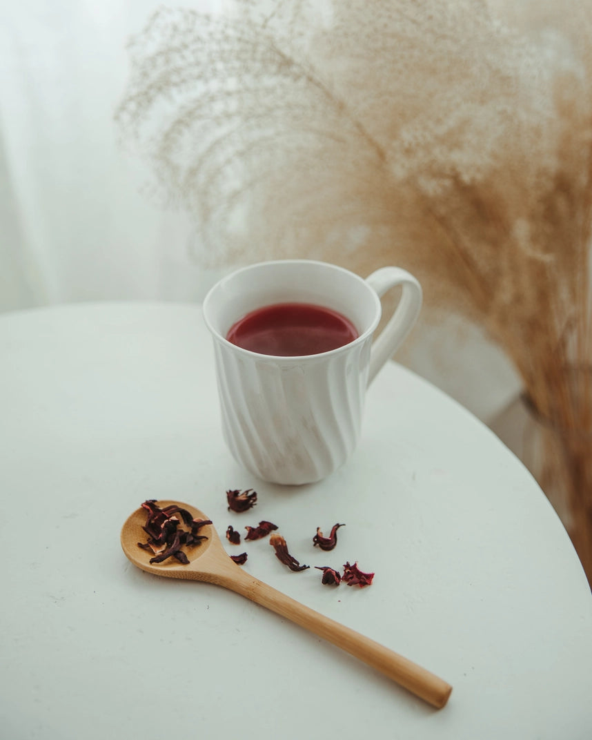 A white cup of red tea on a white table with a wooden spoon and some loose tea sprinkled near it. a plant in background