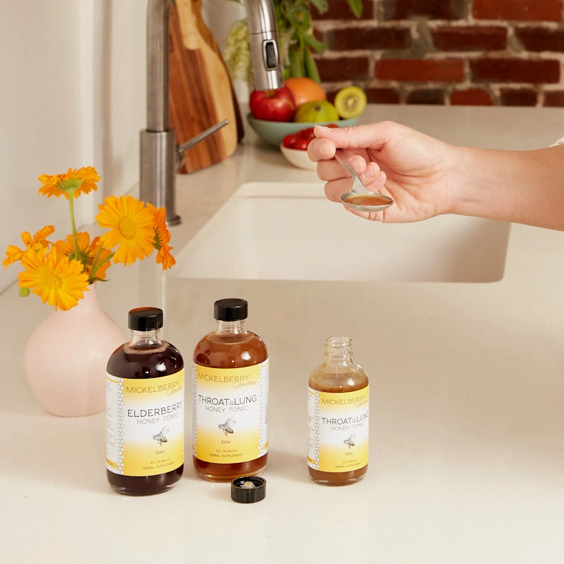 2 8 oz and one 4 oz bottle of tonic one is open with it's lid on a counter. There's a hand holding a spoonful of the tonic and a sink in the background with flowers ans fruit on the counter