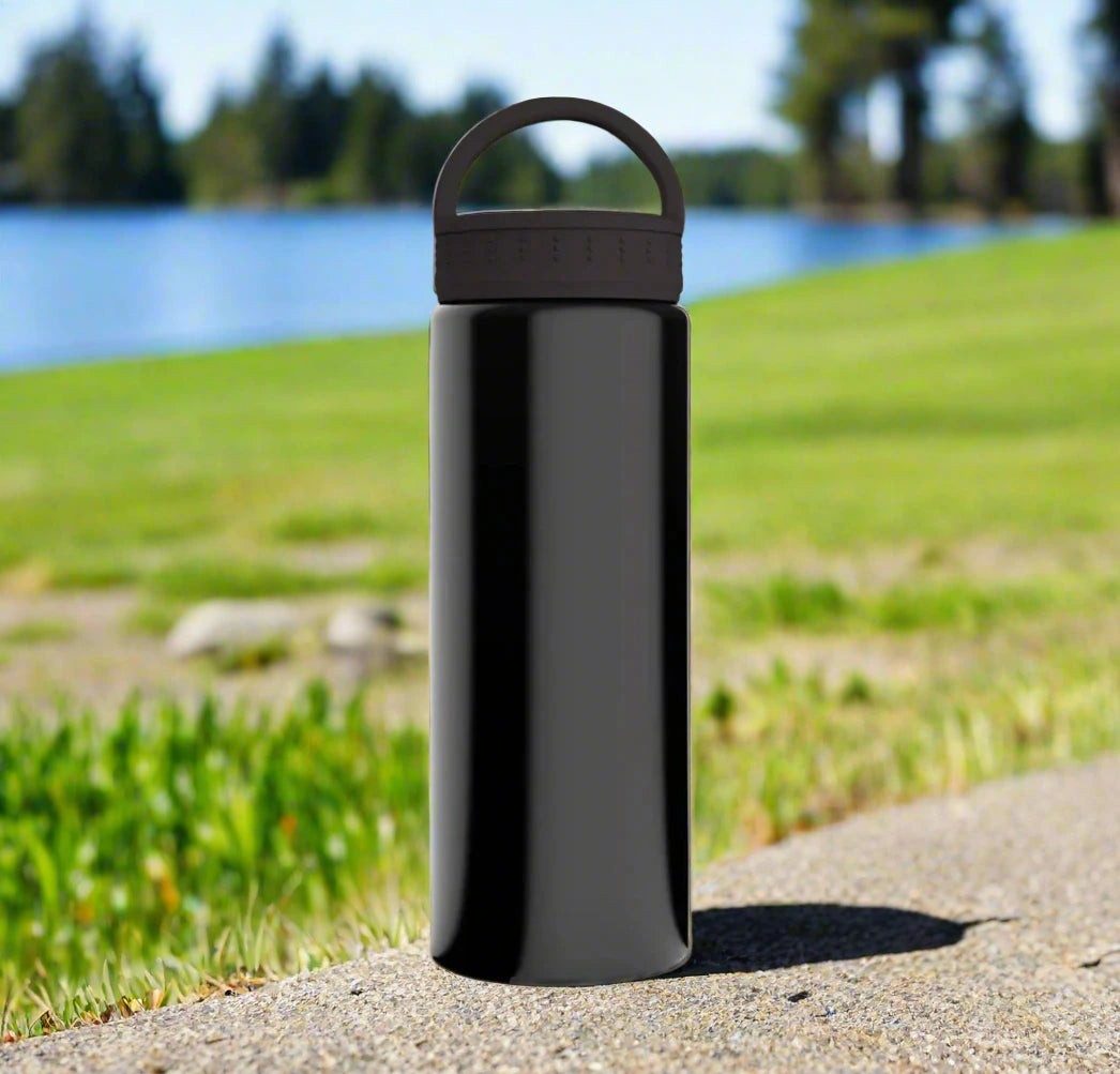 A sleek black water bottle sitting near a tranquil lake, surrounded by lush grass and nature.